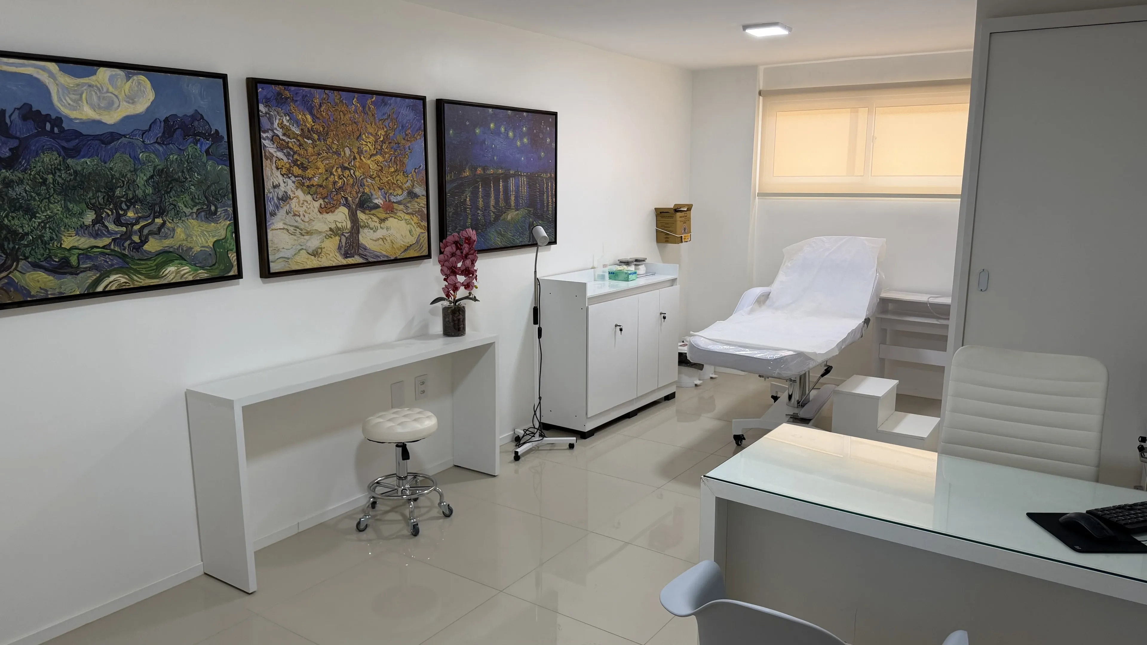 Consultório com maca, armário branco, mesa de atendimento e reproduções de pinturas de Van Gogh