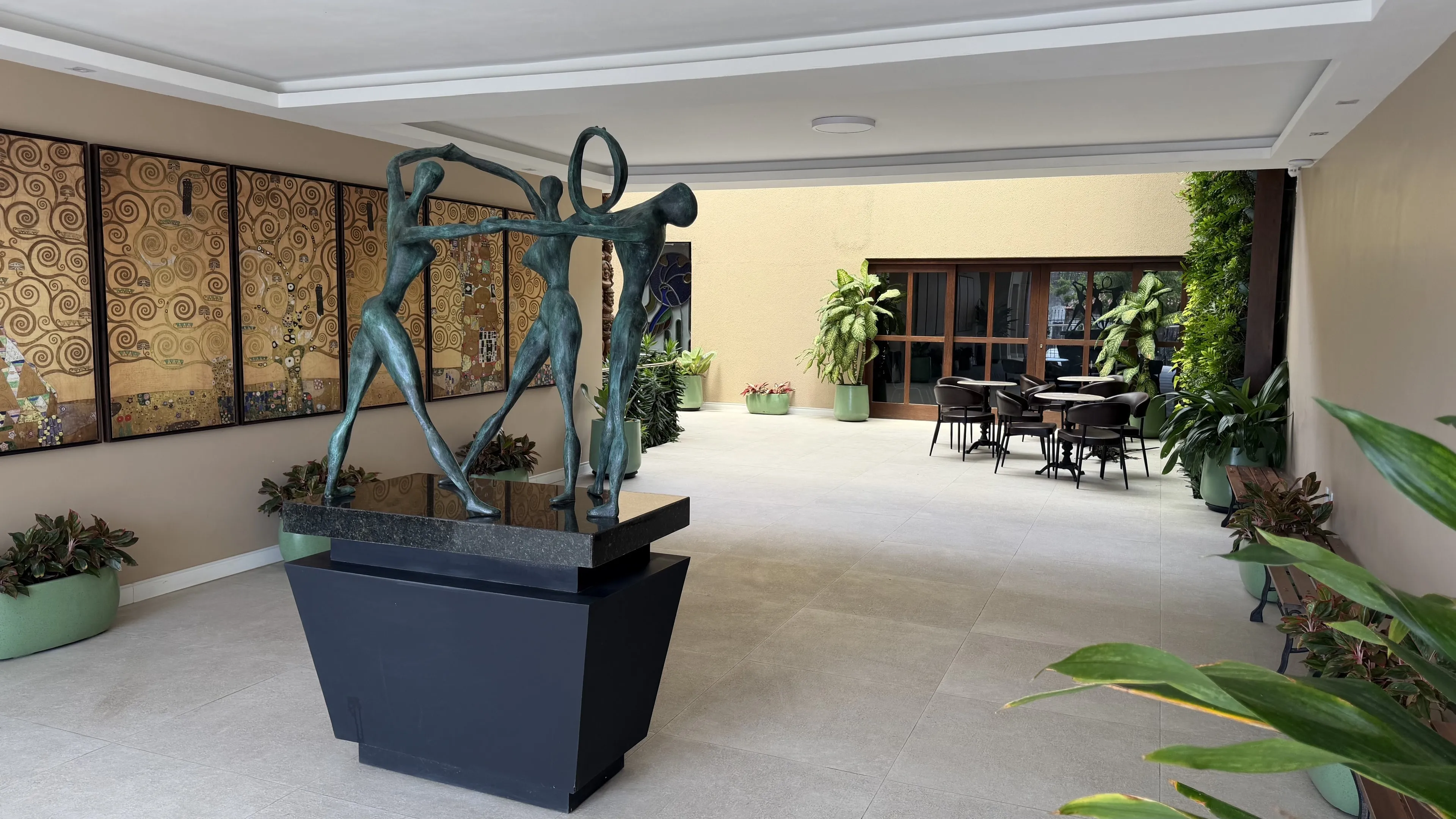 Hall com escultura em bronze de figuras em roda, painéis dourados na parede, plantas e mesas ao fundo