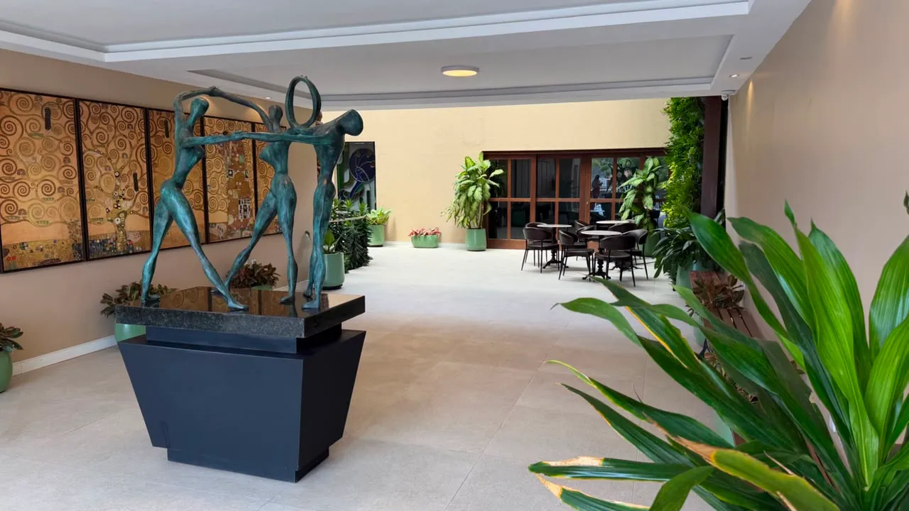 Hall interno com escultura em bronze de figuras em roda, painéis dourados, plantas e mesas ao fundo