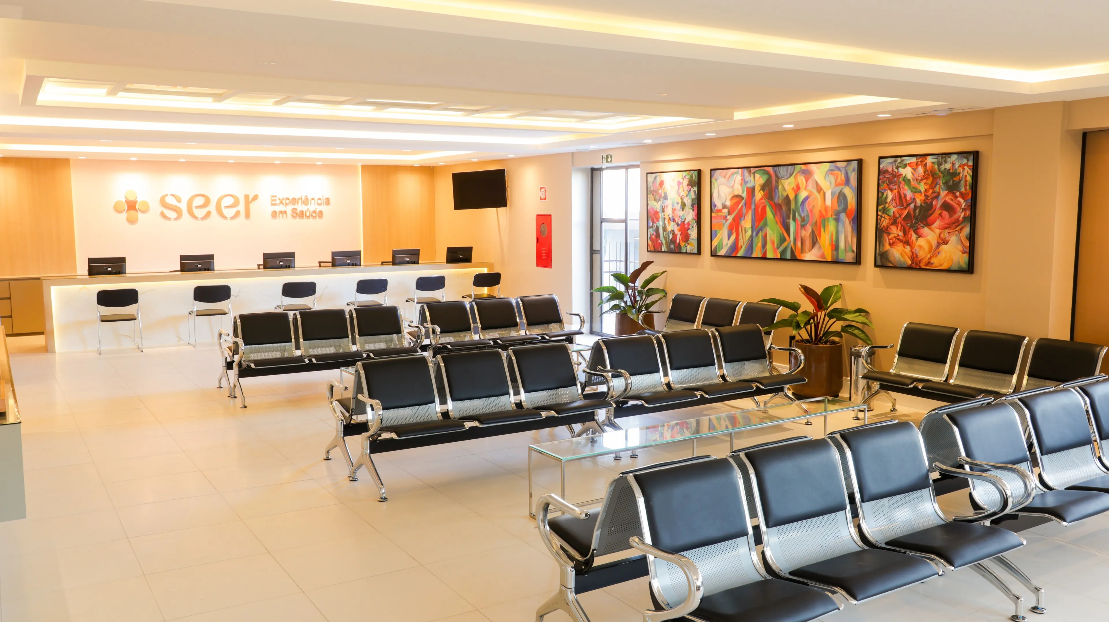 Recepção principal da SEER Saúde com balcão de atendimento, letreiro “seer Experiência em Saúde”, longarinas e quadros coloridos