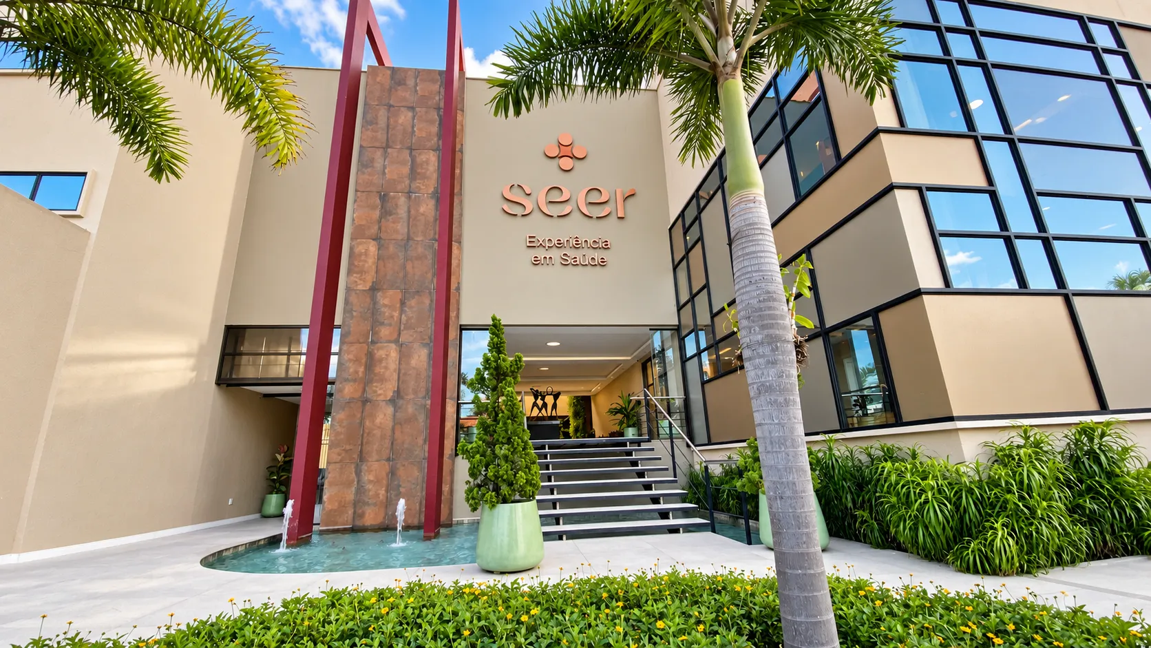 Entrada do edifício da SEER Saúde, com letreiro, espelho d’água, pilares vermelhos e jardim com palmeiras